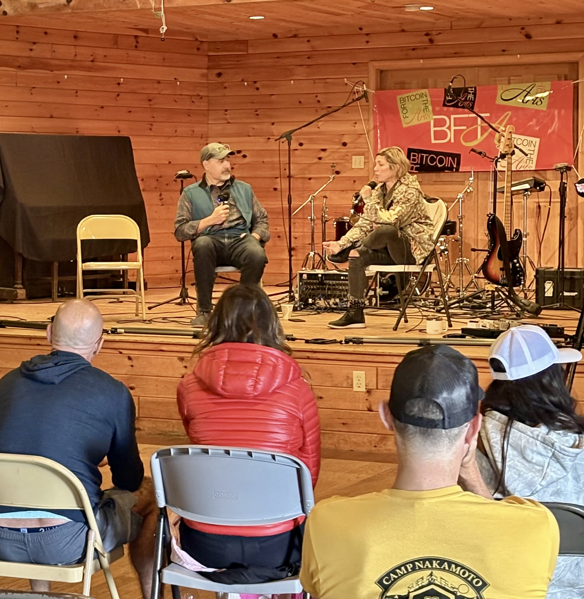A stage discussion at Camp Nakamoto with the audience watching, the Bitcoin for the Arts logo visible on the backdrop